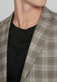 Sivý károvaný blazer s hnedými akcentmi, textúrovaná tkanina a čierny tričko s okrúhlym výstrihom pod ním, viditeľné na výstrihu.