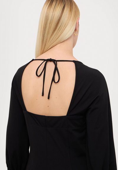 Robe noire à manches longues, dotée d'un large dos ouvert maintenu par un lien. Tissu lisse avec une silhouette structurée.