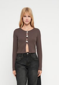 Νεαρή γυναίκα που φορά cropped μακρυμάνικη καφέ μπλούζα με μεγάλα ασημένια κουμπιά και μαύρο τζιν ψηλόμεσο, στέκεται μπροστά σε απλό λευκό φόντο.