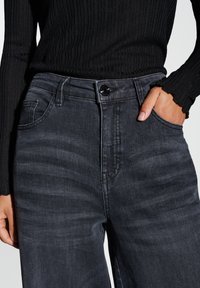 Haut noir à manches longues côtelé associé à un jean taille haute en denim gris foncé. Le jean présente une fermeture à bouton et des poches latérales.