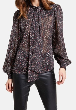 Mujer con blusa negra transparente con pequeño estampado floral rojo y blanco y lazo en el cuello, combinada con pantalones de cuero negros.