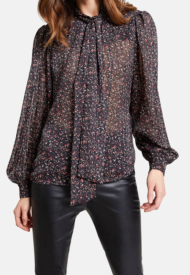 Femme portant un chemisier noir transparent avec un petit imprimé floral rouge et blanc et un col à nouer, associé à un pantalon en cuir noir.