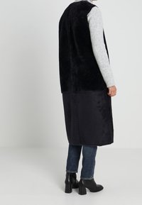 Černá vesta z umělé kožešiny s dlouhým střihem, nošená přes světle šedý žebrovaný svetr, doplněná tmavými džínami a botami na širokém podpatku.