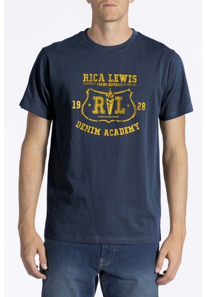 Homme portant un t-shirt bleu marine avec un texte jaune "Rica Lewis Denim Academy 1928" et un emblème, assorti à un jean bleu.