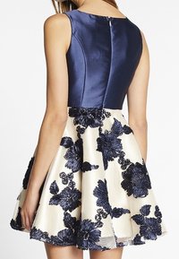 Marinblå satinöverdel med en dold dragkedja, som leder till en krämfärgad kjol prydd med mörk blommönsterbroderi, vilket skapar en lager-effekt.