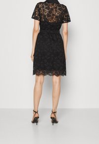 Robe en dentelle noire avec des manches courtes, motif floral, taille cintrée, ourlet festonné et dos transparent. Portée avec des talons noirs à lanières.