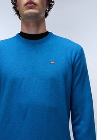 Maglione blu a maglia con scollatura rotonda, caratterizzato da un piccolo dettaglio di bandiera ricamata sul petto. Il tessuto ha una trama liscia.