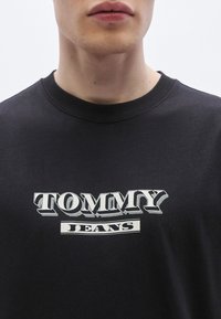 Fekete pamutpóló kerek nyakkivágással, fehér "TOMMY" logóval és alatta "JEANS" felirattal, mindkettő vastag, stilizált betűtípussal.
