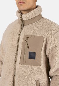 Chaqueta de forro polar beige con cuello alto, cierre frontal con cremallera y un bolsillo en el pecho confeccionado en un material contrastante, con un pequeño parche de logo.