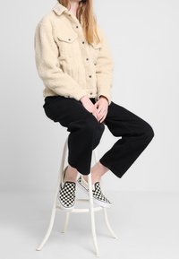 Veste beige en fausse fourrure avec poches avant, pantalon noir à jambe large et chaussures à enfiler noir et blanc à carreaux, assise sur un tabouret blanc.
