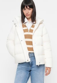 Chaqueta acolchada blanca con capucha desmontable, cierre de cremallera y detalles en plata, sobre un suéter de punto a rayas en tonos marrón y crema, combinado con jeans azules.