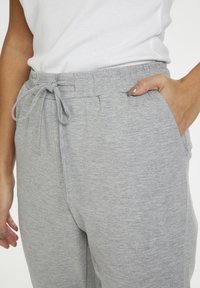 Persoon in lichtgrijze joggingsbroek met een elastische tailleband en trekkoord, hand in de zak, en een wit shirt dat is ingestopt.