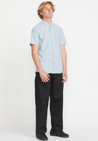 Chemise à manches courtes bleu clair avec un bouton et une poche, associée à un pantalon noir et des baskets foncées, se tenant devant un fond blanc.