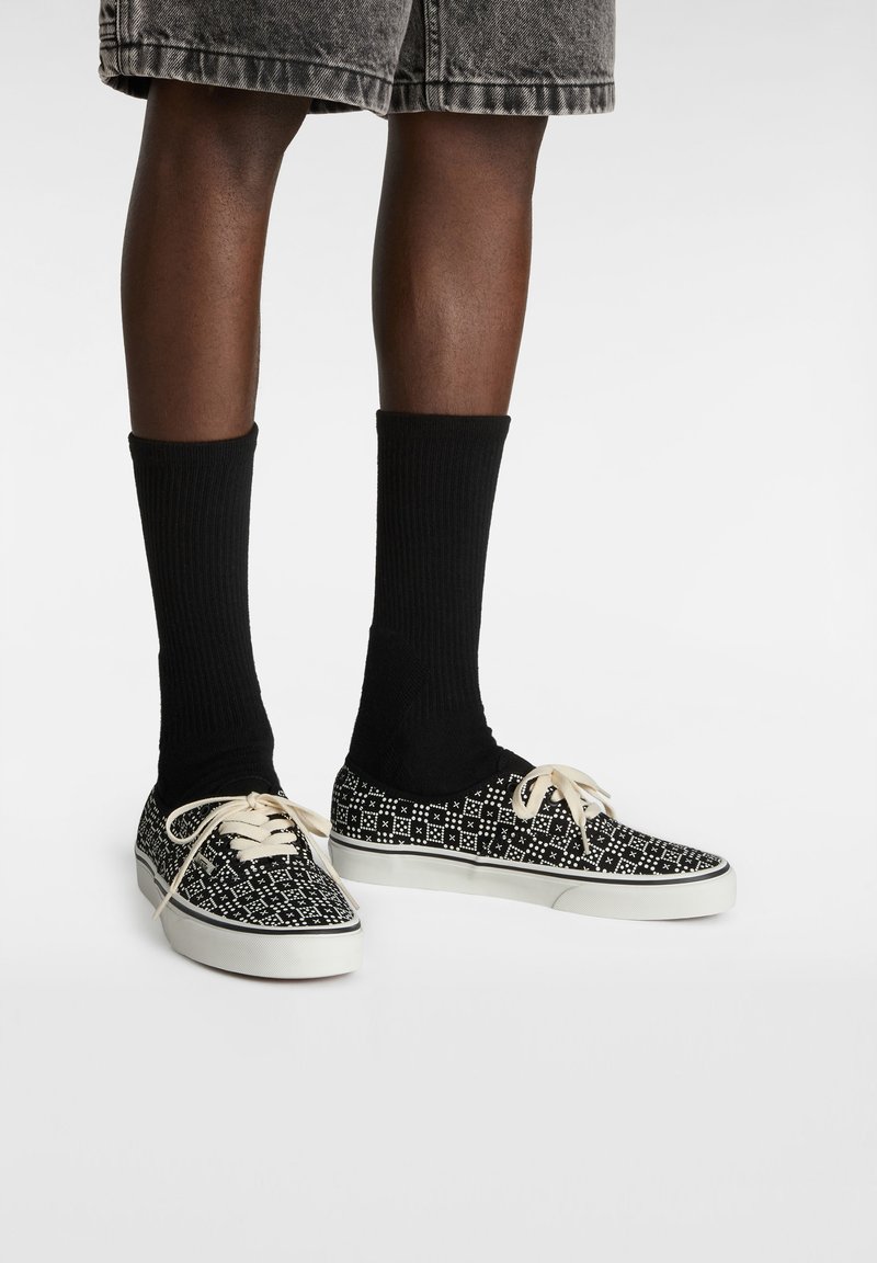 Zapatillas de lona en blanco y negro con un patrón, con cordones crema, usadas con calcetas negras hasta la rodilla. Diseño de perfil bajo con una suela blanca.