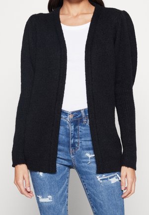 Zwarte chunky gebreide cardigan met een open voorkant, geribbelde textuur en lange mouwen. Gecombineerd met versleten blauwe jeans en een wit topje.