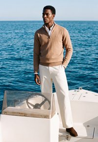 Homme portant un pull beige et un pantalon blanc debout sur un bateau, une main dans la poche, océan en arrière-plan sous un ciel clair.