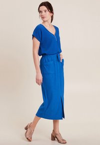 Robe midi bleu côtelé avec manches courtes, décolleté en V, poches latérales et fente avant, associée à des sandales à talons beiges.