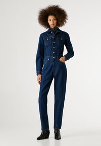 Denim overal v tmavomodrej farbe, s golierom, predným zapínaním na gombíky a náprsníkovými vreckami. Úzke nohavice a dlhé rukávy dopĺňajú dizajn.