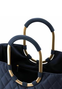 Bolso de tote acolchado en color navy con asas de metal dorado, que presentan empuñaduras azules acanaladas. El interior incluye un bolsillo con cremallera para un almacenamiento seguro.