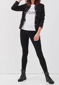 Woman wearing black buttoned jacket, white graphic t-shirt, black skinny jeans, and black lace-up ankle boots standing against white background.