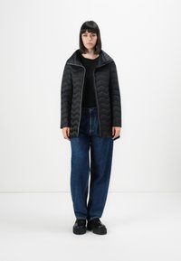 Jonge vrouw in een zwarte gewatteerde jas, zwart shirt, losse blauwe spijkerbroek en zwarte platform schoenen, staande tegen een witte achtergrond.