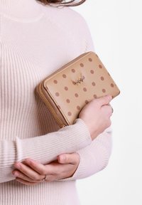 Tan polka dot wallet with a gold logo, featuring a textured exterior and a zip closure, held against a light sweater.