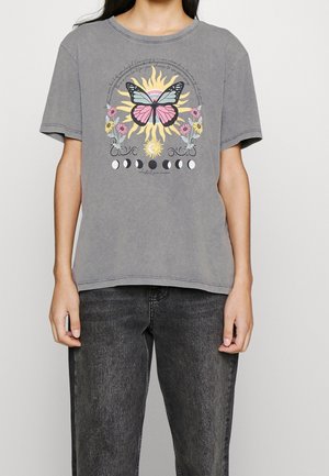 Osoba nosząca szary t-shirt z kolorowym graficznym wzorem przedstawiającym motyla, słońce, kwiaty i fazy księżyca z przodu.