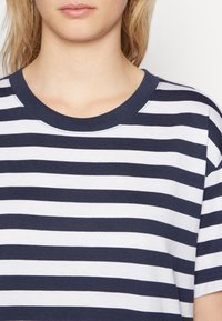 Merin ja valkoisin raidoittein varustettu t-paita, jossa on pyöreä pääntie ja lyhyet hihat, valmistettu pehmeästä puuvillakankaasta. Kauluksessa näkyvä ompeluyksityiskohta.