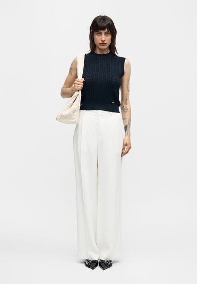 Γυναίκα με σκούρα μαλλιά που φοράει αμάνικη μπλε πλεκτή μπλούζα, λευκό παντελόνι με ευρύ πόδι, μαύρα μυτερά παπούτσια και κρατάει μια κρεμ υφή τσάντα στον ώμο της.