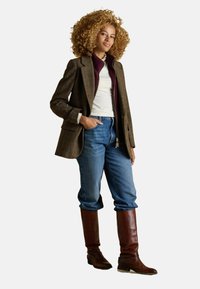 Braun kariertes Sakko über einem burgunderfarbenen Reißverschluss, weißes Hemd, blaue Jeans und braune knielange Lederschuhe. Lockige Haare und entspannte Haltung.