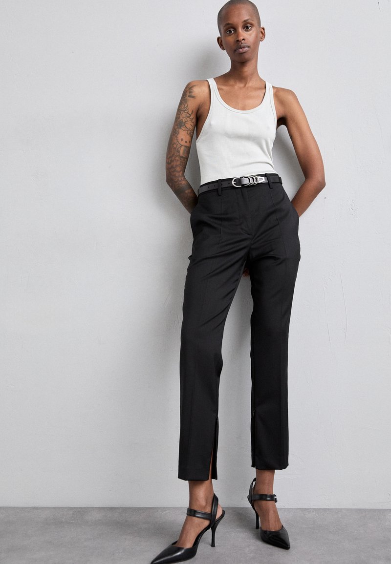 Black tailored trousers with side slits, paired with a white ribbed tank top and black pointed heels, featuring a silver belt with circular accents.