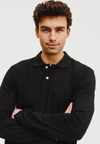 Czarna, długowieczna koszula polo z guzikowym kołnierzykiem, z subtelnym logo marki i gładką teksturą, noszona przez osobę z założonymi rękami.