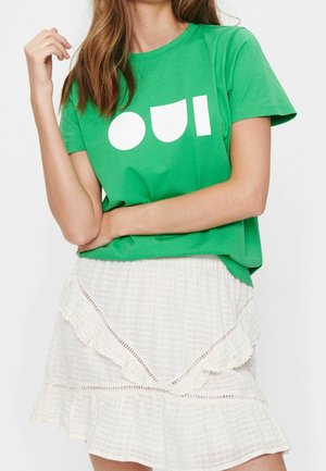 Femme portant un t-shirt vert avec des formes géométriques blanches et une jupe blanche à volants avec un tissu texturé et une bordure en dentelle.
