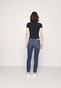 Marinblus med korta ärmar och små röda detaljer, tillsammans med kroppsnära blå jeans och vita sneakers, sedd bakifrån.