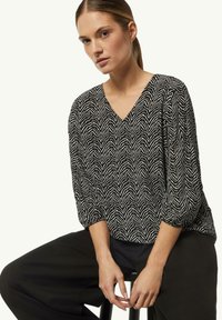 Schwarz-weiß gemusterte Bluse mit V-Ausschnitt, 3/4-Ärmeln und strukturiertem Stoff im Zickzackdesign. Kombiniert mit lockeren schwarzen Hosen.