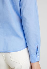 Camisetta azzurra chiara a manica lunga con taglio dritto, polsini abbottonati, texture morbida e orlo leggermente curvo, abbinata a pantaloni bianchi.