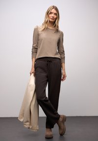 Beiges langärmliges Oberteil mit gepufften Schultern, dunkelbraune, locker sitzende Hose mit Kordelzug, hält eine cremefarbene Jacke und trägt braune Stiefel.