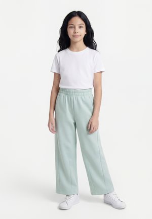 Fille aux longs cheveux noirs portant un t-shirt blanc à manches courtes, un pantalon large vert clair et des baskets blanches, debout devant un fond blanc.