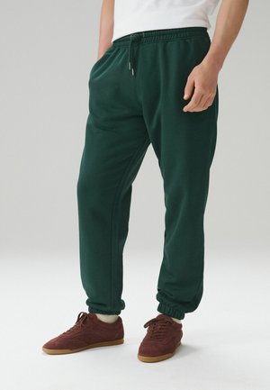 Mann trägt dunkelgrüne Jogginghosen mit elastischen Bündchen und kastanienbraune Sneakers mit Gummisohlen, steht vor einem einfarbigen hellen Hintergrund.