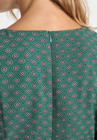 Vihreä kuvioitu paita, jossa on oktaalimuotoilu mustilla, valkoisilla ja punaisilla yksityiskohdilla. Takana vetoketjukiinnitys ja sileä pinta.