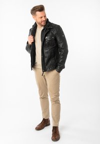 Zwarte leren jas met een fleece kraag, voorzakken, beige shirt, licht beige broek en bruine veterschoenen.