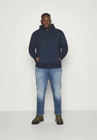 Ενήλικας άνδρας στέκεται, φορώντας μια σκούρα μπλε φούτερ, ανοιχτό μπλε σκισμένα τζιν και ελιόχρωμες μπότες, μπροστά σε απλό φωτεινό φόντο.