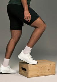 Chaussures de sport blanches posées sur une boîte en bois, associées à un short noir et un haut vert. La chaussure présente une semelle texturée et des lacets.