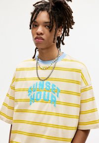 Striped cotton t-shirt in cream, yellow, and blue, featuring "SUNSET HOUR" stitched in blue. Includes layered beaded and metal necklaces.