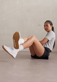 White athletic shoes with gum soles, worn with white crew socks. The outfit includes a gray t-shirt and black shorts, on a light floor.