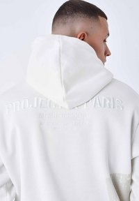 Sudadera blanca con capucha y texto en relieve en la parte posterior. Presenta una textura suave y un suave degradado de color cerca del dobladillo.