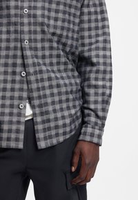 Pitkähihainen napillinen paita, jossa on harmaa-musta ruudullinen kuvio, valmistettu kevyestä kankaasta, jossa on rintatasku ja kaareva helma.