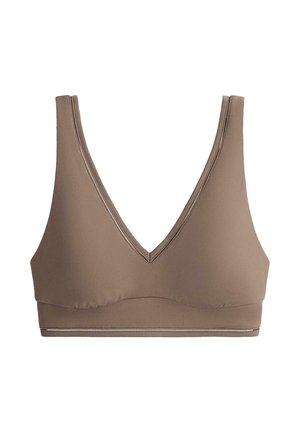 Femme portant un soutien-gorge de sport marron assorti et des leggings taille haute, debout devant un fond clair uni, regardant devant elle.