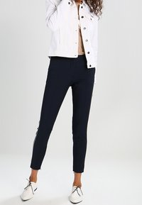 Marina blå skräddarsydda byxor med vita sidostänger, tillverkade av mjukt material, med slim fit och ankellängd, i kombination med en vit denimjacka.