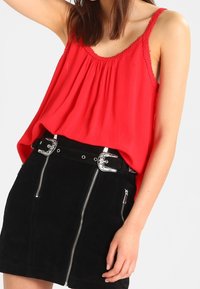 Haut sans manches rouge avec un col froncé et une bordure en dentelle, associé à une mini-jupe en suède noire dotée de fermetures éclair argentées et de boucles décoratives.